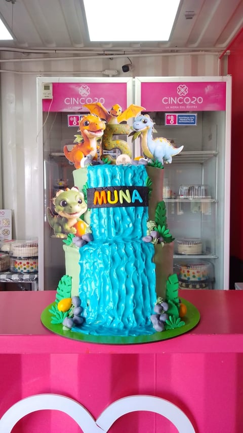 Three-tiered turquoise cake with colorful animal figurines on top and MUNA text, displayed on pink table at event