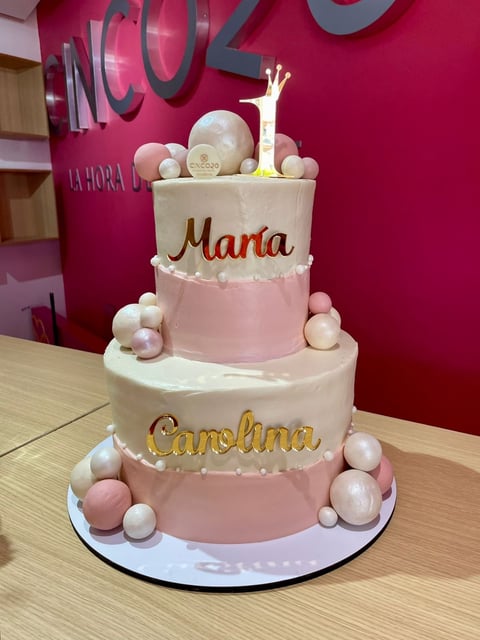 Four-tiered pink and white celebration cake decorated with pearls, featuring names Maria and Carolina, displayed in a bakery setting