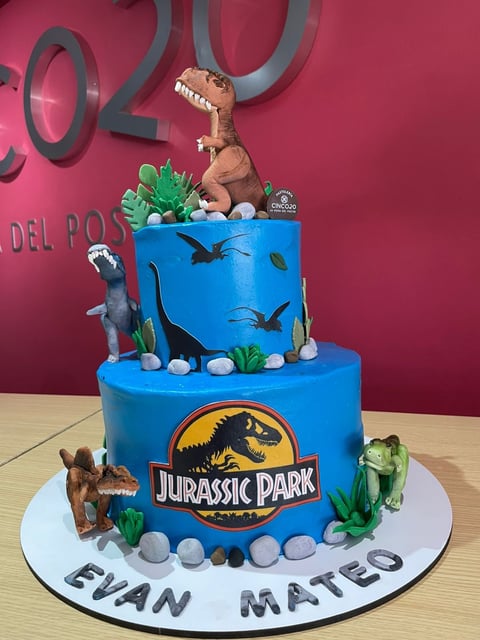 Two-tier blue Jurassic Park themed birthday cake with dinosaur figurines, green plants, and personalized name base.