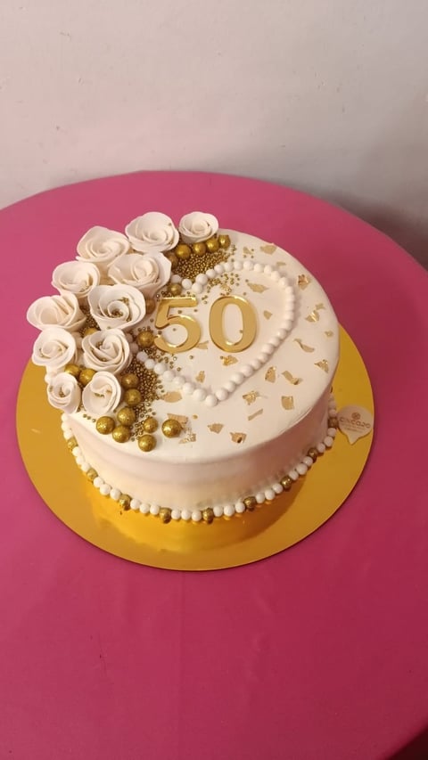 Elegant 50th birthday cake with cream frosting, cream roses, gold 50 number, pearl beads, and gold accents on yellow cake board against pink background