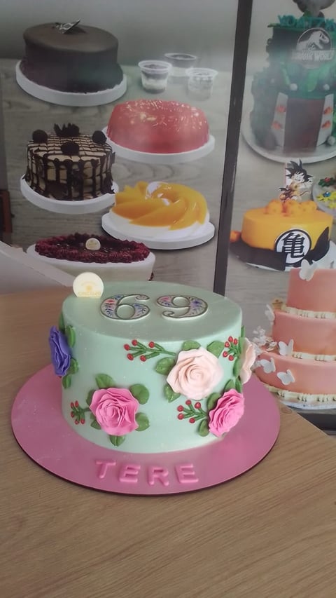 Mint green fondant cake with pink roses and leaves on pink base, displayed in bakery window with other decorated cakes