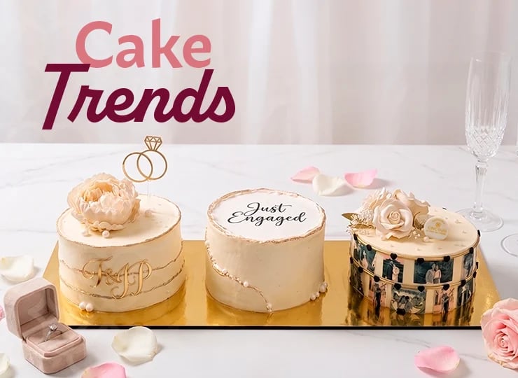 Three elegant engagement cakes displayed on a gold tray with pink roses and ring decor against a Cake Trends backdrop