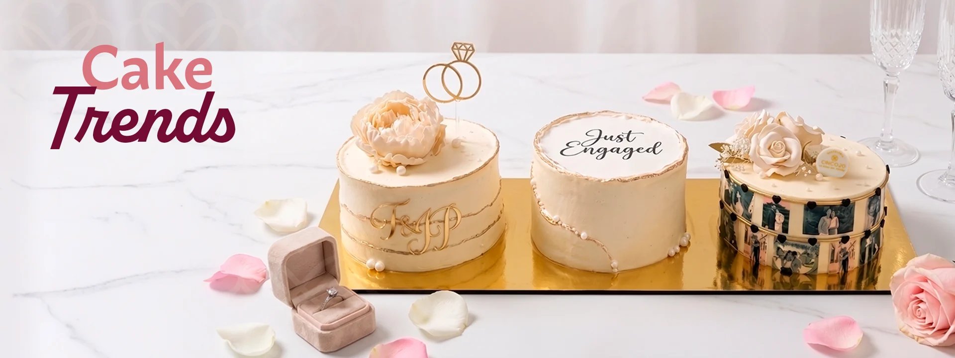 Three elegant cakes displayed on a gold tray with cake toppers, decorated with pink petals and champagne flutes.