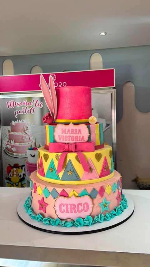 Colorful multi-tiered circus-themed birthday cake with pink, yellow, and blue decorations, featuring feathers and text reading MARIA VICTORIA and CIRCO