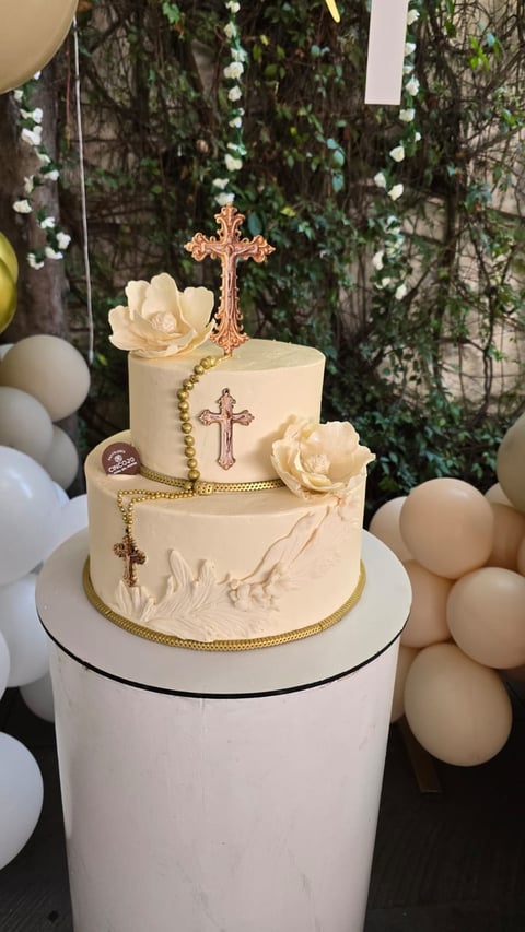 Two-tiered cream cake with gold cross cake topper, decorative crosses, and ivory roses surrounded by white and beige balloons and greenery